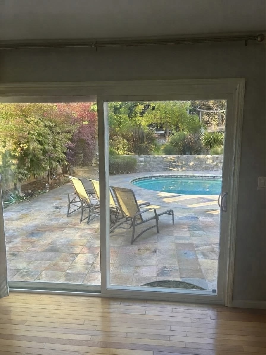 View through a sliding glass door to a stone patio, lounge chairs, and a swimming pool