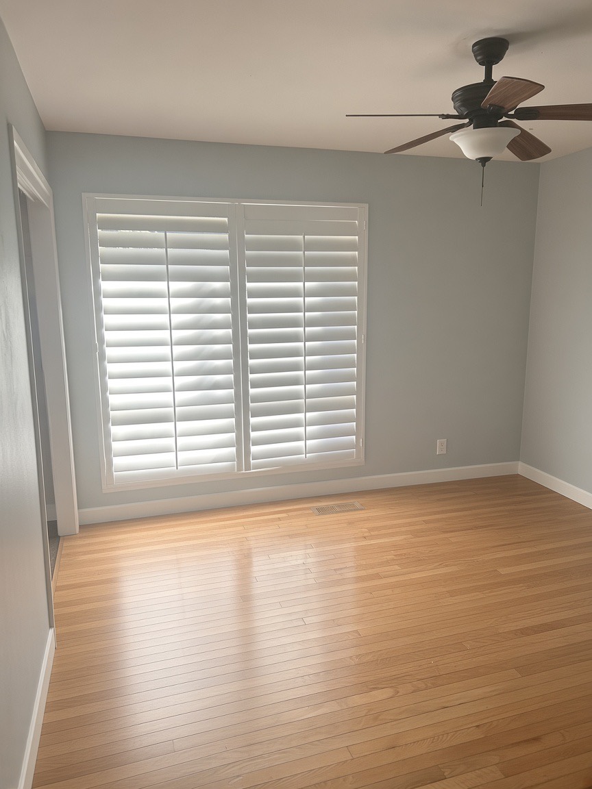 Bedroom with pale blue walls, hardwood floors, white plantation shutters, and a ceiling fan
